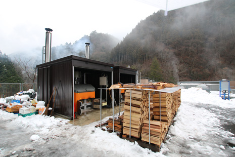 雪景色に白い煙をはきだす薪ボイラーと、近くに積まれた、たくさんの薪木の風景